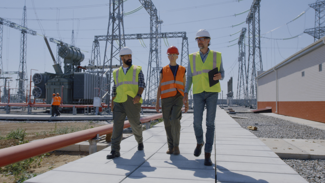 Tracking shot of male worker in helmet and vest walking on path and demonstrating modern power plant to group of engineers